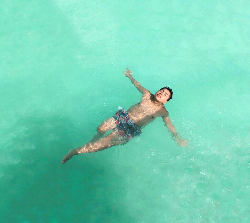 Person floating on their back in clear turquoise water seen from above, arms relaxed, eyes closed — the weightless calm of a float session