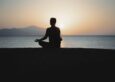 Person meditating in lotus pose silhouetted against a calm sea at sunset